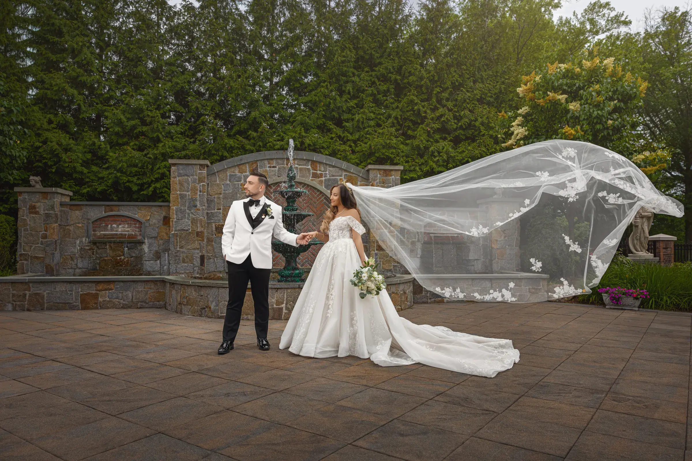 Bride and groom by a garden fountain with flying veil, by George Pompilio