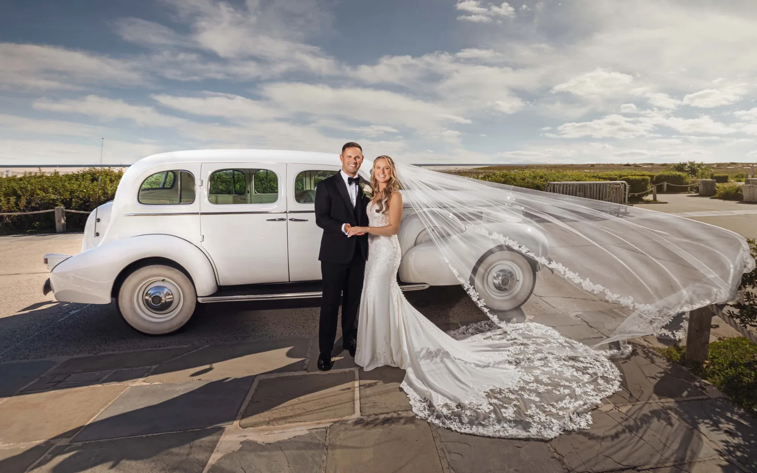 Allison and Luke beside a vintage white car as her veil catches the wind at Gatsby On The Ocean &mdash; George Pompilio Photography