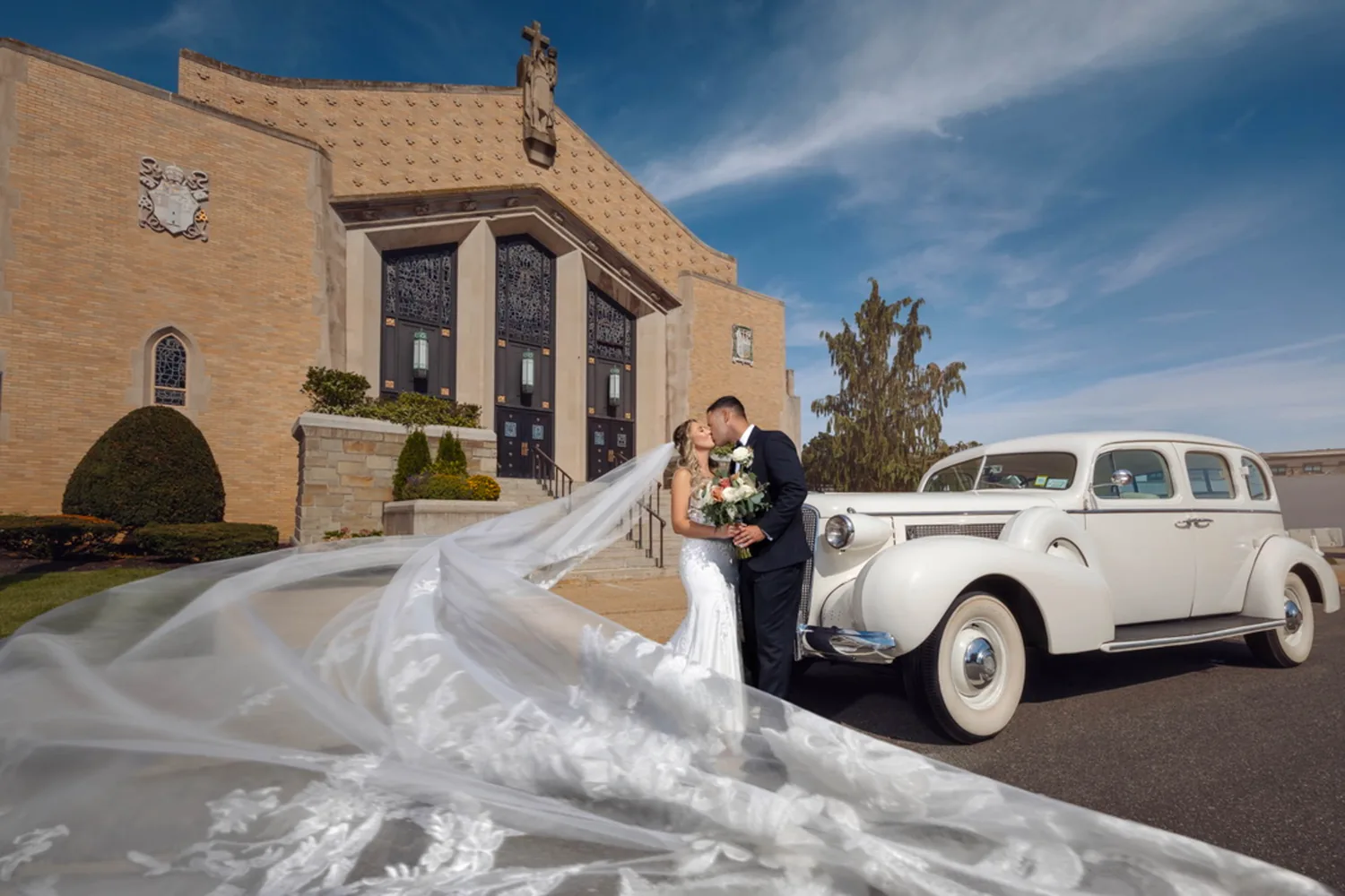 Allison and Luke kissing outside The Church of St. Christopher beside a vintage white car, her veil in the wind &mdash; George Pompilio Photography